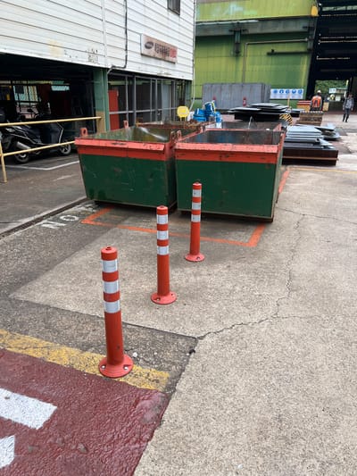 This image depicts an outdoor industrial area in Geoje-si, South Korea. In the foreground, two large, green and red industrial dumpsters are positioned on a concrete surface marked with orange lines and white lettering. Three orange and white striped traffic cones are placed in front of the dumpsters. To the left, a portion of a building with a white, corrugated metal exterior is visible, featuring a sign with Korean text and the Samsung logo. A black scooter is parked under an overhang of the building. In the background, a large green industrial building is present, with stacks of metal plates and industrial equipment visible in front of it. Two individuals, dressed in work attire, are walking in the background near the metal stacks. The scene appears to be a functional area within a manufacturing or processing plant.