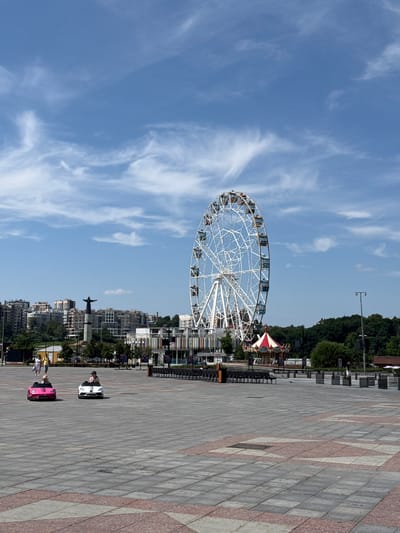 The image depicts a large, paved public square in Cheboksary, Russia, under a bright, partly cloudy sky. In the background, a prominent white Ferris wheel stands tall, with a carousel partially visible nearby. Beyond the amusement rides, a cityscape of multi-story buildings stretches across the horizon, with a statue of a person with outstretched arms positioned atop a pedestal in the left portion of the background. The foreground of the square is occupied by two small, electric ride-on cars: a pink one on the left with a child driver, and a white one on the right, also with a child driver. A few scattered individuals are visible in the middle ground, including a couple walking together and a person standing near the carousel. The paving stones of the square are in various shades of gray and feature decorative reddish-pink geometric patterns. The overall scene suggests a leisurely day at a park or recreational area within the city.