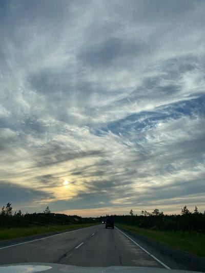 The image depicts a road scene with a dark-colored van traveling away from the viewer on a highway. The sky is filled with wispy, elongated clouds, indicating a potentially overcast or twilight condition, with the sun visible as a bright orb to the left of the frame. The road appears to be asphalt with white lane markings. The surrounding landscape consists of low-lying vegetation and trees, suggesting a rural or sparsely populated area. The vantage point is from inside a vehicle, as indicated by the reflection on the lower portion of the image. There are no discernible geographical landmarks that would definitively place this scene in Lempino, Russia, or any other specific city.