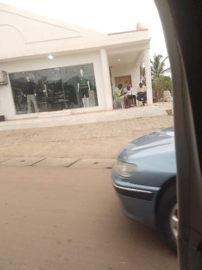 The image shows a scene in New Karu, Nigeria, taken from inside a car looking out. Three men are seated on the sidewalk outside a storefront. One man is wearing a green shirt and shorts, another is wearing a brown shirt and pants, and the third is wearing a blue shirt and pants. They are all looking at something off-camera. The storefront is white and has a large window with a mannequin displayed inside. There are trees to the right of the storefront. The sidewalk is paved with cement and there is a concrete curb separating it from the street. The street is dirt and has a drainage ditch running along the edge. The car that the image is taken from is blue and is parked on the side of the road, facing the storefront. The view is looking forward from the inside of the car, and the car is in the foreground, partially obstructing the view of the scene. The image has a low resolution and the colors are slightly muted. The weather appears to be overcast. The scene is located in New Karu, Nigeria, at the coordinates 8.991 latitude and 7.57 longitude.