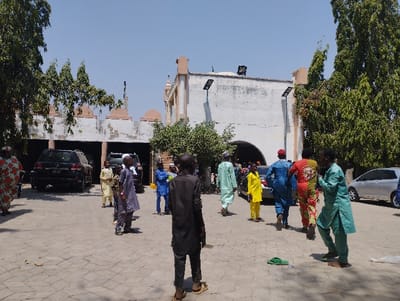 The image shows a group of people standing and walking in a courtyard in front of a white building with an arched doorway. The people are mostly men and boys, and they are wearing traditional clothing. The courtyard is paved with concrete tiles and there are several cars parked in the background. The building has a white facade with a large arched doorway and a flat roof. The courtyard is located in front of a white building with an arched doorway. The building has a flat roof and a white facade. There are several cars parked in the background. The building is located in Kaltungo, Nigeria. The coordinates of the location are 9.816 latitude and 11.315 longitude. There is a large tree on the left side of the frame and another large tree on the right side of the frame.  The sky is blue and clear. The sun is shining and there are shadows on the ground. The image appears to have been taken on a bright, sunny day. There are also several trees around the building. The image appears to show people attending a religious gathering.