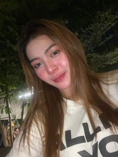 The image is a close-up, selfie-style photograph taken at night, likely outdoors, with the subject positioned in the foreground. The subject is a young woman with long, light brown hair that falls around her face. She is wearing a white long-sleeved shirt with a large, black graphic print that appears to read "ALL NYC". Her face is illuminated, and she has makeup on, including blush and lipstick. The background is dark and out of focus, but appears to be an outdoor setting with foliage and some artificial lighting, possibly string lights or spotlights. In the blurred background on the left, there are some structures and possibly other people, suggesting a public or semi-public area like a restaurant, bar, or garden. Without further context, it is impossible to definitively place the image within Bandar Lampung, Indonesia.