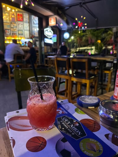 A close-up, high-angle shot shows a red, frothy drink in a textured glass with a black straw, placed on a table in what appears to be a sports bar. The drink is positioned in the foreground, obscuring some of the surrounding objects. The table surface is a light-colored wood. To the left of the drink, a menu or coaster features graphics of sports balls, including a basketball and a football, with the text "VIET NAM" visible. Behind the drink, a blue menu or promotional item is partially visible with text that reads "QUY NHON SPORTS BAR" and "VIET NAM". To the right of the drink, a silver ashtray rests on the table, containing what looks like cigarette butts. The background is blurred but suggests an indoor/outdoor bar setting with wooden chairs and tables, neon lights, and people seated in the distance. The overall ambiance is dimly lit with colorful lighting from neon signs and decorative lights. The establishment is identified as "Quy Nhon Sports Bar" in South Quy Nhon Ward, Vietnam, based on the text present.