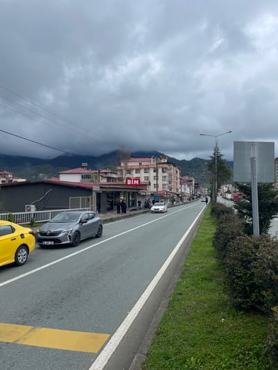 The image shows a street scene in Kemalpaşa, Turkey. The street is lined with buildings on the left, including a BIM store, and a green space on the right. Several people are walking on the sidewalk in front of the buildings. Two cars, one yellow and one gray, are parked on the side of the road. Mountains are visible in the background, partially obscured by clouds. The sky is overcast. A street lamp and a signpost are visible along the roadside.