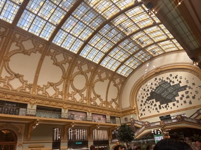 This image captures the magnificent interior of a grand, multi-story building, likely a historic structure repurposed as a modern commercial space, consistent with locations like the Stadsfeestzaal in Antwerp, Belgium. The scene is dominated by a vast, arched glass and metal ceiling that bathes the space in bright natural light, suggesting it is daytime with clear weather.
The architecture is opulent, featuring cream-colored walls adorned with extensive gold leaf or painted intricate baroque or Belle Époque style decorative patterns and moldings. Several levels of balconies with ornate black and gold balustrades wrap around the central hall, showcasing various retail establishments.
On the ground floor and upper levels, signs for shops and eateries are visible. Prominently displayed is "WEEKDAY," a fashion retailer, on the lower left. To the right, a large sign reads "ZEST," indicating a food or beverage outlet, below a grand, sweeping staircase with marble steps and decorative railings. Further up the staircase, another sign identifies "INNO," a department store. On one of the upper balconies, a banner partially shows the text "SHELFIE STORAGE."
On the large wall to the right, a striking piece of modern art or decoration is mounted, consisting of numerous dark, square panels arranged in a scattered, geometric pattern that converges in the center. A few blurred figures of people are visible in the lower foreground, suggesting general activity like shopping or strolling within this public space. Potted trees or large plants add touches of greenery to the luxurious environment. The overall impression is one of a bustling, elegant indoor plaza.