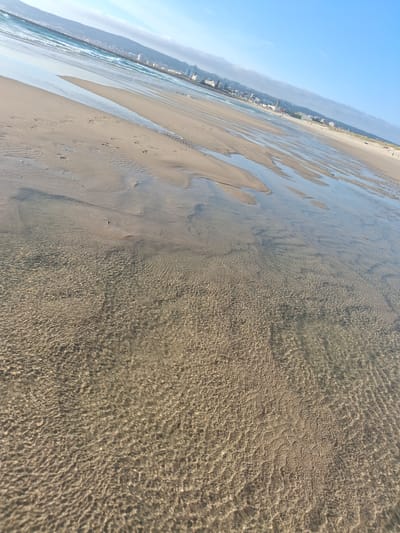 This photograph shows a beach scene with clear shallow water covering the sand, creating intricate patterns of light and shadow on the seabed. In the background is the urban shoreline of Figueira da Foz, Portugal, with buildings visible along the coast under a clear blue sky. The tide appears to be low, revealing a wide expanse of the beach. There are no people visible in the immediate foreground, and the overall impression is one of stillness and natural beauty contrasted against the backdrop of the city.