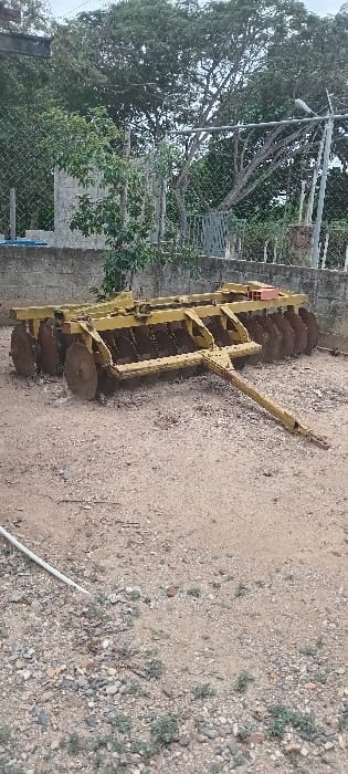 An agricultural disc harrow is positioned on dry, light brown soil mixed with pebbles and sparse green vegetation in Cujisal, Venezuela. The implement, painted yellow with visible rust on its numerous circular steel discs, faces towards the foreground with its hitch pole extending to the lower right. A white pipe is partially exposed on the ground in the lower-left quadrant. Behind the harrow, a low concrete wall supports a chain-link fence. Beyond the fence, a dense collection of green trees and foliage forms the background, including a large tree prominent in the upper right and a concrete block structure partially visible in the upper-left. A metal gate is discernible through the fence in the mid-right background. No individuals are present.