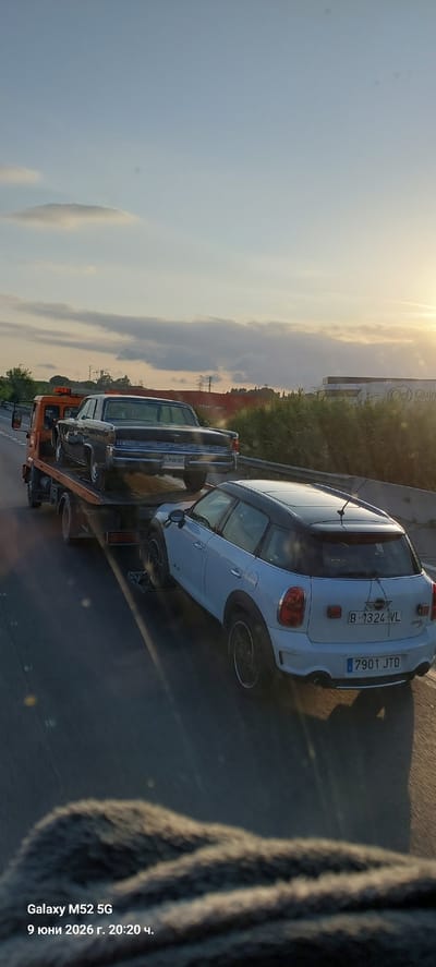 An orange tow truck is carrying a black Lincoln Continental on its flatbed, driving on a highway at sunset. In the foreground, a white Mini Cooper Countryman with a black roof is being towed. The Mini Cooper's license plate reads "B-1324-VL" and a secondary plate shows "7901 JTD". The Lincoln's license plate is partially visible and reads "H2666BDC". The scene appears to be on a highway near industrial buildings with tall power lines in the background. The sky is clear with some clouds, and the sun is low on the horizon. The bottom of the image shows a blurry close-up of a furry texture, likely a seat cover or blanket, with overlaid text indicating the phone model "Galaxy M52 5G" and the date and time "June 9, 2026 20:20". The location is identified as Santa Perpètua de Mogoda, Spain. There are no people visible in the image.