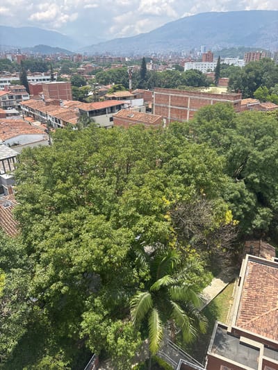 This image depicts an aerial view of a neighborhood in Medellín, Colombia. The foreground is dominated by lush green trees interspersed with buildings, predominantly featuring red-tiled roofs. A multi-story brick building under construction is visible. A cityscape extends into the background, with mountains in the distance. The lighting suggests a bright, sunny day, with some cloud cover.