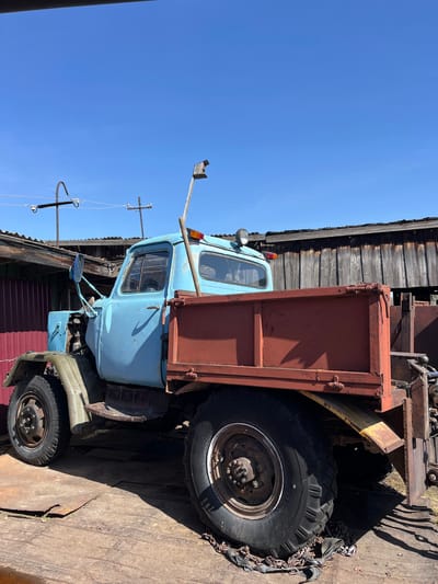 A faded blue and red dump truck sits adjacent to a weathered wooden building with a rusty corrugated metal lower section in Верхняя Уря, Russia. The truck's tires are thick and worn, with one tire wrapped in chains. The sky above is clear blue. Utility poles with wires are also visible. The ground appears to be covered with wooden planks. The truck appears old and in a state of disrepair.