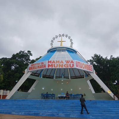 The image shows the Grotto of the Virgin Mary in Moshi, Tanzania. The Grotto is a structure with a blue and white roof, adorned with a ring of stars and a cross on top. Banners with red lettering reading "GROTTO YA BIRIKA MARIA" and "MAMA WA MUNGU UTUOMBEE" are displayed on the structure.
People are present in the scene. One man is walking up the blue stairs, while others are sitting on chairs inside the Grotto. Trees are visible in the background, adding to the environment. The sky is overcast.