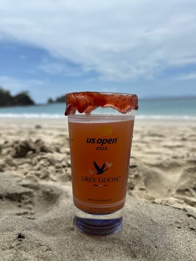 A plastic cup containing an orange-colored beverage, with its rim coated in a dark red, granular substance, rests partially embedded in light-colored sand on a beach in Sardinal, Costa Rica. The cup is branded with "US OPEN 2025," the "GREY GOOSE" logo featuring three flying geese, and the hashtag "#HONEYDEUCE." Small text at the bottom edge states "SIP RESPONSIBLY." The immediate foreground consists of sandy terrain with visible ripples and texture. The midground features a sandy beach leading to a calm, turquoise-blue ocean. In the background, several small, tree-covered rocky islets emerge from the water on the left, while the ocean extends to the horizon on the right. The sky is blue with scattered white clouds. No individuals or active subjects are present in the scene.