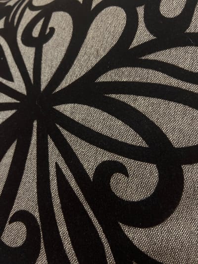The image shows a close-up view of a patterned fabric. The background fabric appears to be a woven material, likely a linen or similar blend, with a greyish-tan color. A bold, swirling, floral-like design in black velvet is superimposed on the background fabric. The design features thick, curved lines that form petal-like shapes and decorative swirls. The composition is asymmetrical, with the design extending beyond the frame on multiple sides. The texture of both the woven background and the velvet design is visible. There is no information available to suggest that this image was taken in Cherliany, Ukraine, or any specific location. The absence of people, interactions, or any environmental clues in the close-up image makes it impossible to determine the setting.