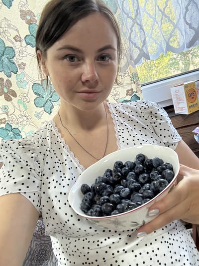 A woman is holding a white bowl filled with blueberries. The bowl is tilted towards the camera. She is wearing a white top with black polka dots and lace trim. A thin necklace is visible on her neck. Her fingernails are painted with a light, reflective polish. In the background, there is a window with a patterned curtain, a partially visible box with Chinese characters, and an outdoor scene. The location appears to be in the city of Малая Рукавицкая, Russia.