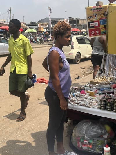 This image captures a bustling street scene in Ablekuma, Ghana, under bright daylight. In the foreground, a dark-skinned woman stands in profile, facing right, with short, curly, light brown hair. She wears a light purple sleeveless tank top and dark leggings, with light-colored slip-on sandals. Her left hand rests near her hip. To her right, a market stall features a display of silver and black beaded necklaces, small packaged goods, and stacked circular metal tins on a white surface. Below the stall, a large clear plastic bag contains various colorful items, next to a red-capped plastic bottle. A yellow sign attached to the stall advertises "MTN MOBILE MONEY ALL NETWORKS," showing logos for MTN, Airtel, Tigo, and Telecel Cash, along with transfer rates.

To the left of the woman, a dark-skinned man walks leftward in profile. He wears a bright lime-green short-sleeved patterned shirt, dark green shorts, and brown leather sandals, carrying a dark plastic bag in his left hand. Partially visible behind the market stall is another person wearing a light grey tank top and black shorts.

In the mid-ground, a white two-door hatchback is parked. The street surface is a mix of asphalt and packed dirt, with scattered debris. The background reveals a busy urban environment with additional market stalls under colorful awnings (green, red), various pedestrians, and multiple buildings constructed from concrete and corrugated metal. Utility poles and overhead wires are also present.