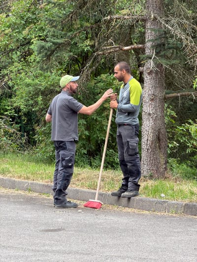 In Leudelange, Luxembourg, two men are standing near a large tree and a curb. The man on the left, wearing a grey polo shirt, grey cargo pants, and a green cap, has a beard and is gesturing with his left hand towards the man on the right. The man on the right is wearing a grey and lime green long-sleeved shirt and grey cargo pants. He is holding a broom with a red brush and a wooden handle. They appear to be engaged in a conversation. The environment is lush with green foliage and grass, with a paved surface in the foreground.