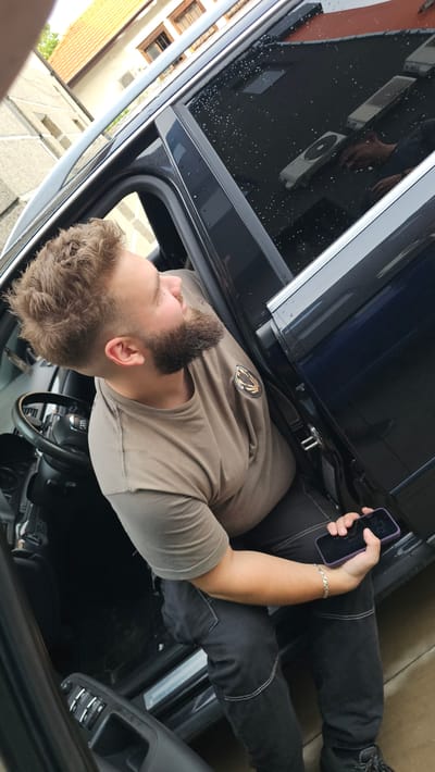 A Caucasian male with a beard and short, styled blonde hair is partially seated in the driver's side of a dark-colored SUV. He is wearing a tan t-shirt with a circular emblem on the left chest and dark jeans with reinforced stitching. His left arm rests on the steering wheel, and his right hand is holding a purple smartphone. The car door is open, revealing the interior which includes the steering wheel, dashboard, and window controls on the door panel. The window is wet with visible water droplets and reflects an external scene that includes a white air conditioning unit and a partial reflection of the man himself. The vehicle appears to be parked outdoors, possibly in a driveway or street, with residential buildings and a tiled roof visible in the background. The scene is set in Karlovo, Bulgaria.