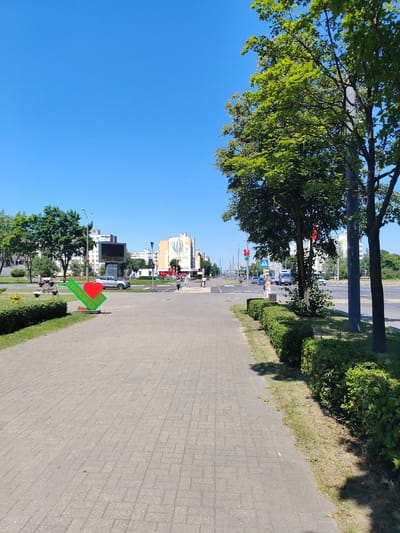 An eye-level, medium shot captures a sunny day in Salihorsk, Belarus. A paved walkway runs diagonally from the lower left to the upper right of the frame, bordered by manicured green bushes. To the left of the walkway, a prominent sign featuring a red heart and green lettering that reads "СОЛИГОРСК МЫ" (Salihorsk We) is visible. Across the street, buildings are visible, including one with a large digital billboard. Trees with vibrant green leaves line the street and sidewalks. A crosswalk is present, with pedestrians walking on the opposite sidewalk. Cars are also present on the multi-lane road. The sky is a clear, bright blue.