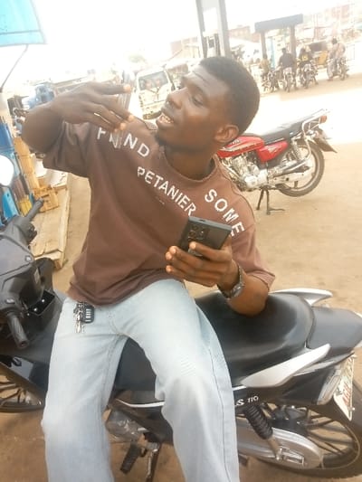 A man sits astride a black and silver scooter. He wears a brown shirt with white lettering and light-wash jeans. In his right hand, he holds a black cell phone. Keys dangle from his jeans. The scooter model appears to be a "JS 110." A red motorcycle sits parked behind him. In the background, more motorcycles are visible along with a white truck. The setting appears to be an outdoor public area, possibly a street or parking lot.