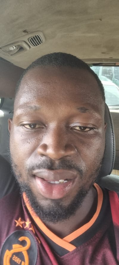 The image is a close-up selfie of a Black man with short black hair and a trimmed beard. He is wearing a maroon and orange soccer jersey featuring three stars and the text "2002". The man is inside a vehicle, likely a car or truck, as evidenced by the vehicle interior, including air vents, visible in the background. Based on the image information, it's impossible to confirm the location in Satellite Town, Nigeria. There are no other people, subjects, interactions, or environments visible.
