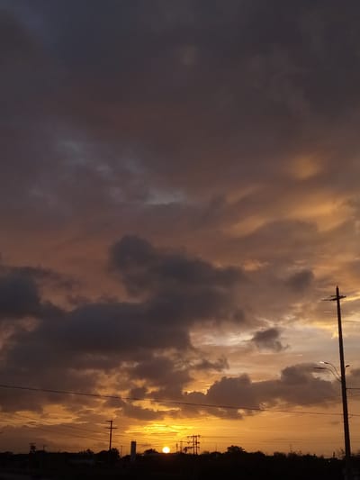 A sunset is captured in El Dátil, Venezuela, with the sun partially obscured by clouds. The sky displays a gradient of colors, from deep purple and grey in the upper portion to bright orange and yellow near the horizon. Silhouetted against the vibrant sky are utility poles with electrical wires stretching across the frame, and the dark outline of trees and buildings along the horizon.