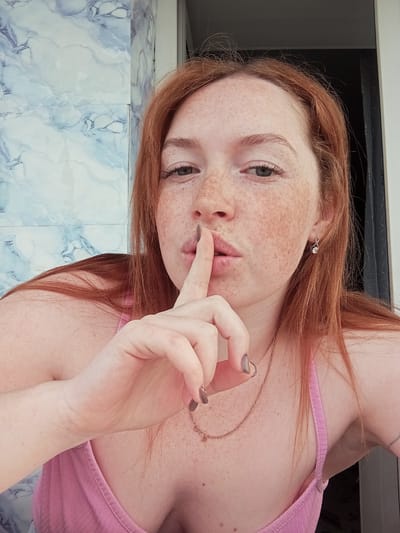 A fair-skinned woman with red hair and numerous freckles is posing with her finger to her lips. She is wearing a pink top and a delicate gold necklace. An earring is visible in her left ear. The background consists of white and blue patterned tile on the wall and a dark gray surface, possibly a window frame or a door. The location is unknown.
