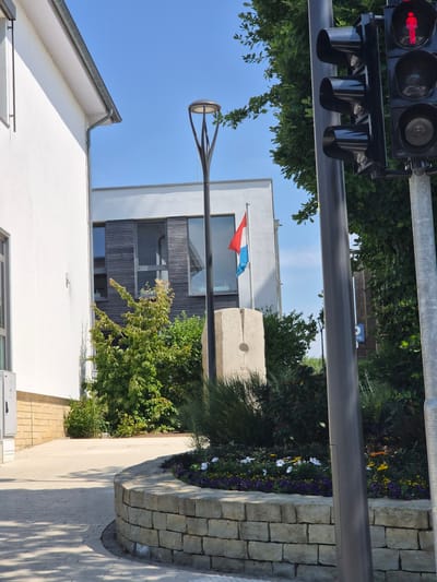 This image captures an outdoor scene on a clear, sunny day, likely in the afternoon due to the strong shadows cast on the pavement. The composition is oriented vertically.
In the foreground, a modern white building with a gray-tiled roof is visible on the left. To the right of the building, a pathway paved with light-colored stones leads towards a landscaped area. This area features a low stone wall constructed from irregular blocks, and a variety of plants, including green bushes and flowering plants with white and purple blossoms.
In the middle ground, a modern building with white upper walls and a dark wooden-paneled section is prominent. A flagpole with the flag of Luxembourg (red, white, and light blue horizontal stripes) is mounted in front of this building. Adjacent to the flagpole is a rough, stone monument or marker with etched lines and a circular indentation, suggesting it might be a sundial or a boundary marker. A streetlamp with a distinctive modern design stands near this monument.
In the background, more lush green trees and a bright blue sky are visible. A traffic signal with three black housings is mounted on a metal pole at the top left of the frame. The red arrow light is illuminated, indicating traffic direction.
The overall impression is one of a well-maintained public space, possibly in a town or village in Luxembourg, given the flag and the presence of a traffic signal. The scene evokes a sense of calm and pleasant weather. There is no visible text other than on the traffic signal, which reads "TRAFFIC".