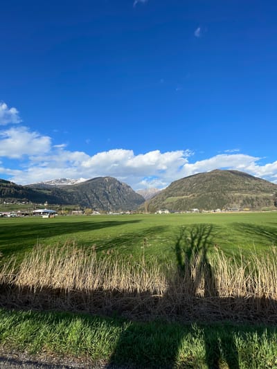 The image depicts a landscape in Sterzing-Vipiteno, Italy. A clear blue sky with scattered white clouds dominates the left side of the frame. Two mountain ridges ascend upwards from the bottom right, one partially covered in snow. A strip of green grass, transitioning to a patch of dry grass at the bottom, extends into the field. Shadows, possibly from trees, are cast on the grass. Buildings and a possible airfield are visible along the base of the mountain range.