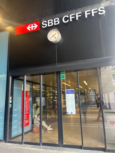 The image displays the entrance of a building, identified as a Swiss Federal Railways (SBB CFF FFS) facility, likely a train station in Geneva, Switzerland. The building features a prominent black facade with "SBB CFF FFS" written in large white capital letters. A classic Swiss railway clock, characterized by its distinctive red second hand, is mounted on the black wall, indicating the time is approximately 11:55. Below the text and clock, a red SBB logo with directional arrows is visible.
The entrance comprises multiple large glass panels, offering a view into the building's interior. Inside, a person is visible lying on the floor or a low surface, dressed in a light-colored jacket and boots, with a brown bag placed next to them, suggesting they are resting or waiting. Reflections of other individuals, display screens, and architectural elements are noticeable in the glass panels. Further within the entrance, a blue information sign with white pictograms and text, and a large red display board, can be seen. The exterior ground appears to be paved. The lighting is diffused, suggesting either an indoor setting or an overcast sky, without strong shadows.