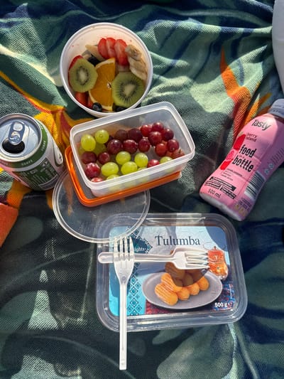 An overhead, eye-level shot captures a picnic spread on a patterned blue and green towel, illuminated by sunlight creating strong shadows. In a white bowl, a colorful assortment of fruit slices includes strawberries, kiwis, oranges, blueberries, and bananas. Adjacent is a clear plastic container holding red and green grapes. A silver can with a green label and the word "SUAVE" is positioned to the left. To the right, a pink bottle labeled "Tasty food in a bottle" and "Berry Island" stands upright. In the foreground, a clear plastic container showcases a serving of golden-brown, syrup-coated pastries labeled "Tulumba" with a 500g weight indication. A white plastic fork rests on top of the Tulumba container. The background is dominated by the textured pattern of the towel. The scene is consistent with a picnic in a public outdoor space.