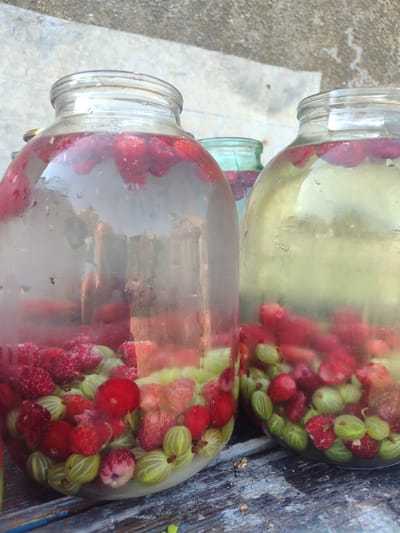 In Novosamarka, Ukraine, three large glass jars filled with raspberries and gooseberries submerged in liquid are placed on a weathered wooden surface. The jar on the left, largest of the three, is partially obscured by condensation and contains a dense collection of red raspberries and green gooseberries. A second, slightly smaller jar to its right contains a similar mix of berries and a clearer, yellowish liquid. Behind these, a smaller, greenish-tinted jar is visible, also containing berries. The background consists of a textured, grey surface, possibly concrete, with a hint of greenery suggesting an outdoor setting. The lighting indicates daytime.