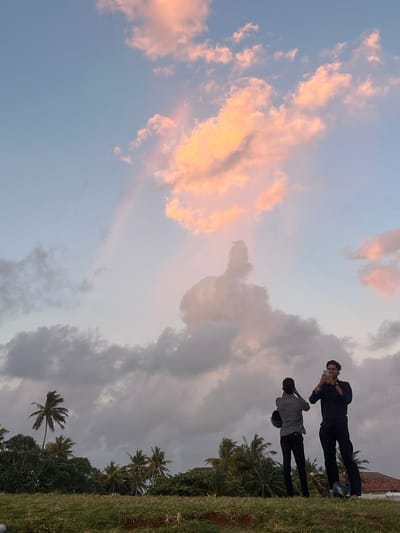This media file captures an outdoor scene with two individuals and a dramatic sky. Two young men are the primary subjects, positioned in the upper right quadrant of the frame. The man in the foreground is wearing a black shirt and black pants, and appears to be taking a photograph with his phone. He is looking down at the device. Behind him and slightly to the left, another man is visible wearing a checkered shirt over black clothing. He is also looking down, possibly at his phone or another device. They are standing on a grassy incline, with palm trees and dense foliage forming the background to their right.
The sky is a prominent feature, filled with a mix of pale blue and soft grey clouds. Towards the upper left, some clouds are illuminated with a warm, peachy-pink hue, suggesting the setting or rising sun. This play of light and color in the clouds creates a visually striking effect, indicative of either dawn or dusk. The overall atmosphere feels serene and contemplative.
The location context provided is Galle, Sri Lanka. This aligns with the presence of palm trees and the tropical feel of the vegetation. The grassy embankment and the hint of a brick structure in the far upper right corner could be indicative of coastal fortifications or a park area. No visible text is present in the media file. The activity appears to be a casual moment of leisure, possibly sightseeing or enjoying the natural beauty of the location.