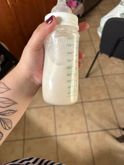 A close-up, vertical, eye-level shot shows a person's left hand holding a baby bottle filled with a foamy, light-colored liquid, likely milk or formula. The bottle is made of clear plastic and has blue measurement markings indicating volume in milliliters and ounces. The liquid level is between the 210 and 240 ml marks. The person's thumb and index finger are visible, with a dark burgundy nail polish on the fingernail of the index finger. A tattoo of outline drawings of leaves is visible on the person's forearm. The background is blurred, showing a tiled floor and what appears to be wooden cabinetry and a dark chair. The image is not specific to Bydgoszcz, Poland.