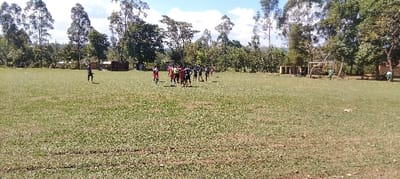 The image shows a grassy field, possibly a soccer pitch, located in Bungoma, Kenya (latitude: 0.554, longitude: 34.774). There are approximately 15 individuals present, all male and young. The individuals are wearing various sports clothing, including red jerseys and shorts. They are engaged in a soccer game, with several people standing together in the middle of the field.  In the background, there are a few trees and a wooden structure, possibly a shed, to the right of the field. The field is mostly empty, and the sky is clear with some light clouds.