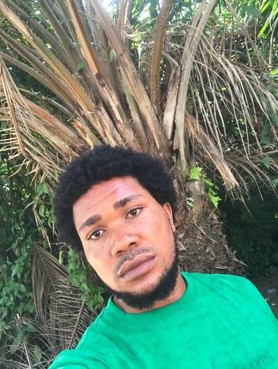 The scene is an outdoor close-up portrait located in Igbologun, Nigeria. The primary subject is an adult male with dark skin, prominent dark, curly hair styled in an afro, a full dark beard, and a mustache. He has full lips and is looking directly into the camera. He is wearing a plain green short-sleeved t-shirt. The background consists of dense tropical vegetation, dominated by the textured trunk and fronds of a large palm tree positioned centrally behind the man. Additional green leafy foliage and some dried palm fronds are visible to the left and right. The scene is illuminated by natural daylight.