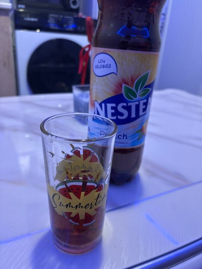 This image captures an indoor scene in Sadovo, Bulgaria, dominated by a bottle of iced tea and a drinking glass. In the immediate foreground, a clear glass stands upright, featuring a decorative design of red watermelon slices and the word "Summertime" in yellow script. A small volume of dark, reddish-brown liquid is present at the bottom of this glass. Behind and slightly to the right of the foreground glass, a large plastic bottle of Nestea Peach flavored iced tea is visible. The bottle's label displays the "NESTEA" logo, "Peach" flavor, and a "LOW CALORIES" speech bubble icon. A thin red ribbon or string is tied around the bottle's neck. The bottle contains a dark, translucent liquid. Further in the background, another blurry, clear glass is partially visible behind the Nestea bottle. The objects are resting on a light-colored, possibly marble or laminate, table surface exhibiting a subtle linear texture. In the deep background, a white household appliance, likely a washing machine, is identifiable by its dark, circular loading door. Above this appliance, a black electronic device, possibly an audio receiver or amplifier, displays several small illuminated blue indicators. The entire scene is bathed in a distinct artificial blue light, casting cool tones and highlighting reflections on the surfaces. No individuals are present.