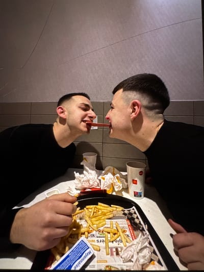 The image captures two young adult males engaged in a playful interaction inside a McDonald's restaurant located in Kumanovo, North Macedonia. Both individuals, with short dark hair and wearing black long-sleeved shirts, are seated opposite each other at a white table, leaning forward with their faces close. Each is holding a single French fry in their mouth, with the tips of the fries meeting in the center between them. Both subjects are smiling.

On the white table, a black plastic tray holds a substantial quantity of golden-brown French fries. Crumpled white paper, likely napkins or wrappers, is interspersed among the fries and on the tray's checkered paper liner. A blue and white packet labeled "Mayonnaise" is visible on the tray, resting over some fries. Two white McDonald's branded paper cups are also present on the table; one is partially obscured on the left, and another, featuring the red "M" logo and red/blue graphics, is visible on the right. The background consists of a light gray or beige wall with horizontal paneling or tiling, indicative of a commercial interior, illuminated by overhead artificial lighting.