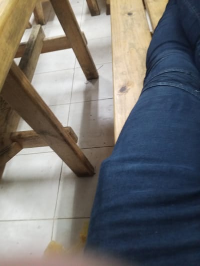 A person's legs, clad in dark blue jeans, are visible, resting on a wooden bench. The bench is made of light-colored, weathered wood planks. To the left, parts of a wooden structure, possibly chairs or tables, are visible. The floor appears to be tiled in a neutral color, with visible grout lines. The overall scene suggests an indoor or covered outdoor setting, possibly in El Valle del Espíritu Santo, Venezuela, given the context provided.