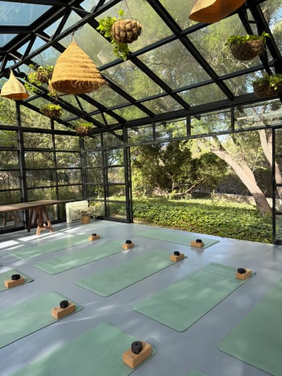 This is a full shot of a yoga studio with a glass roof and walls, allowing for a view of the surrounding greenery. The studio has a grey floor, with multiple green yoga mats laid out, each equipped with two cork yoga blocks and a strap. In the background, there's a wooden table on trestle legs, a white heater, and a basket with dried flowers. The studio is filled with natural light, casting shadows on the floor. The view outside the glass walls shows trees, bushes, and a stone wall, suggesting a natural, possibly rural setting. The overall impression is one of tranquility and connection with nature. The yoga studio is located in Montuïri, Spain.