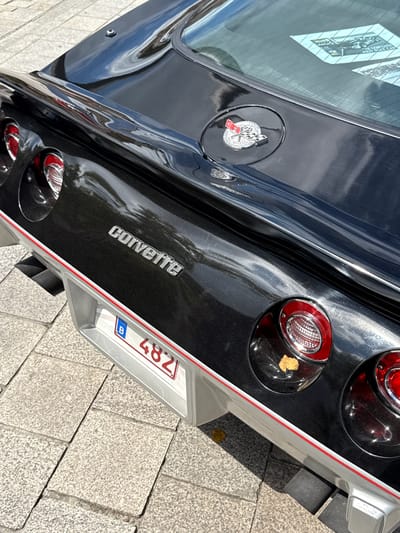 A black Chevrolet Corvette C3 is parked on a cobblestone street in the city of Bruges, Belgium. The rear of the car, including the taillights and the "Corvette" emblem, is visible. A Belgian license plate reading "B 4821" is attached to the rear bumper. The car is clean and appears to be in good condition, with a faint reflection of the sky visible on its polished surface.