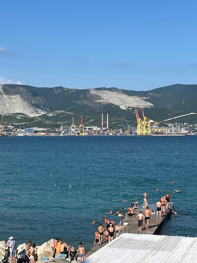 The image depicts a coastal scene on a sunny day. In the foreground, people are gathered on a rocky shoreline and a concrete pier. Some are swimming or wading in the turquoise water, while others are sunbathing, sitting, or standing on the pier and the rocks. Several individuals are interacting on the pier, with some appearing to be preparing to jump into the water.
The middle ground features the expanse of the sea, with a few buoys visible. In the background, a cityscape stretches along the coastline, characterized by industrial facilities with numerous cranes, silos, and other structures. Beyond the city, a range of mountains rises, some appearing to have been quarried, with visible scars on their slopes. The sky is clear and blue. The overall impression is a busy recreational area adjacent to an industrial port. The location is identified as the city of Merchanskoye, Russia.