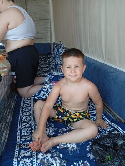 A young boy sits on a blue patterned couch, smiling at the camera. He is shirtless and wearing floral swim trunks. Beside him, a person in a white sports bra and black shorts sits facing away from the camera. The scene appears to be outdoors, possibly in a shaded seating area, with a beige curtain in the background and a tiled wall visible on the left. A black plastic bag lies on the couch to the right of the boy. The image is well-lit, suggesting it was taken during the daytime. The overall mood is relaxed and casual. The location is Teraktagi, Uzbekistan. No visible text is present in the image.