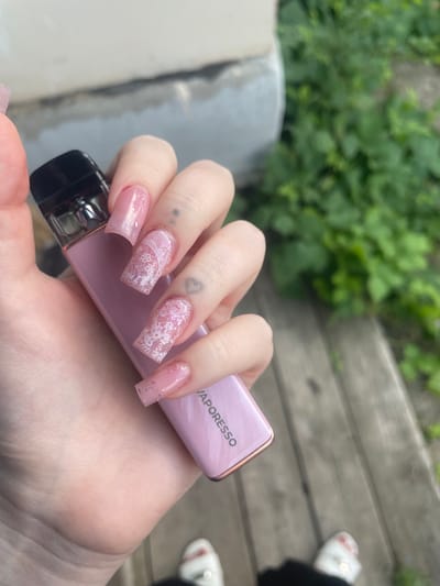 The image shows a close-up of a person's hand with long, pink, square-shaped artificial nails, decorated with white lace patterns and glitter. The hand is holding a pink Vaporesso vape device. The background is slightly blurred, showing greenery, wooden planks, and what appears to be a concrete structure. The lighting suggests it's daytime, possibly outdoors. There are no clear indicators of weather or specific emotions. Visible text on the vape device reads "VAPORESSO". The scene suggests a casual moment, potentially outdoors, given the background elements.