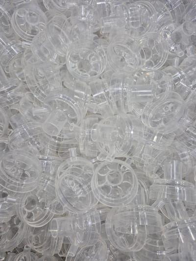 A high-angle, close-up, full-frame shot captures a dense pile of clear plastic medical components, possibly nebulizer cups or similar respiratory accessories, filling the entire frame. The objects are stacked and jumbled together, with many showing a circular base and a tubular protrusion. The plastic is translucent, allowing some light to pass through and creating subtle reflections and shadows. The scene is devoid of any human presence or recognizable environmental context, suggesting an industrial or storage setting. The lighting is even and diffused, highlighting the texture and forms of the plastic parts. No specific location in Pabradė, Lithuania, can be inferred from the image itself.