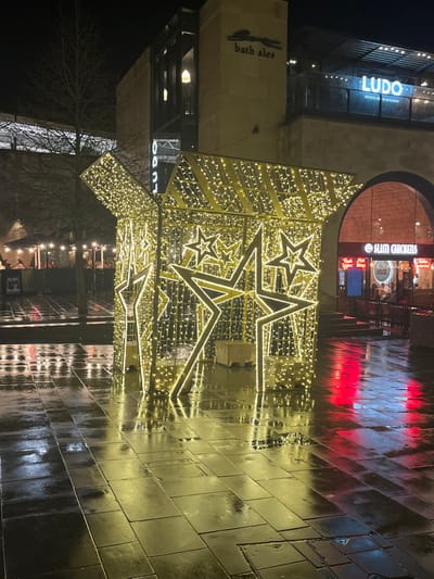 A night scene features a large, lit-up star structure adorned with numerous small lights. The ground appears wet, reflecting the light. Buildings in the background display signs for "Ludo," "bath ales," and "slim chickens." The star structure dominates the foreground, while reflections add depth to the scene. A tree is also visible, adding a natural element amidst the urban setting.