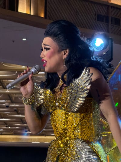 A drag performer, identified as Miss Britney Vegas, is on stage at the J Centre Mall in Cebu City, Philippines, performing in front of an audience. She is holding a silver microphone in her right hand and singing, her mouth open and eyes closed. Vegas is wearing a dazzling gold and silver costume adorned with sequins and iridescent embellishments, featuring prominent wing-like shoulder pieces. Her makeup is dramatic, with bold red lipstick, winged eyeliner, and flushed cheeks. The background shows stage lighting and a wooden ceiling structure.