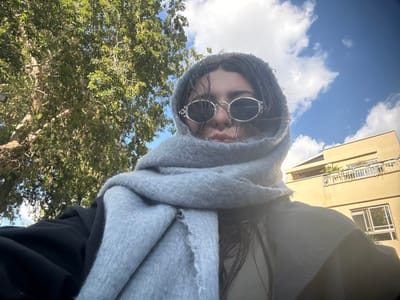 An outdoor, upward-angled photograph depicts an individual in Ramat HaSharon, Israel. The subject, appearing female, is shown wearing a thick, light grey scarf wrapped around her head and neck, partially obscuring her face, and large, round, silver-framed sunglasses covering her eyes. Dark hair is visible around the edges of the scarf and sunglasses. She is dressed in a dark-colored outer garment, consistent with a jacket or coat. The background on the left features a large tree with green foliage and visible branches. On the right, a multi-story, light yellow building with a white-railed balcony and windows is present. The sky above is blue with scattered white clouds, indicating daylight conditions.