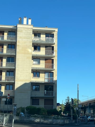 This is a medium-shot, eye-level, exterior photograph taken during daylight. The image showcases a section of a light beige stone apartment building, angled from the right foreground to the left mid-ground. The building features multiple stories with numerous balconies, each equipped with white railings. The balconies have dark-framed windows and some have brown shutters. Atop the building, there are three white chimneys with dark caps. A prominent shadow from the building falls diagonally across the lower left portion of the facade.
In the foreground, a metal fence runs horizontally across the bottom of the frame, with a sidewalk and road beyond. A traffic sign with a red circle and horizontal bar is visible to the left of the building, mounted on a pole. Green bushes and some smaller trees are situated behind the fence and in front of the lower levels of the building.
To the right of the apartment building, in the mid-ground and background, there are more trees, a street, and vehicles. A modern bridge-like structure with a railing and streetlights is visible in the far right background. The sky is a clear, bright blue.
The sign on the pole near the red and white sign appears to read "Mairie Briand" and "Bus," indicating the location is likely near the town hall or a bus stop in L'Haÿ-les-Roses, France. The presence of French street signage and the architectural style of the building are consistent with this location.