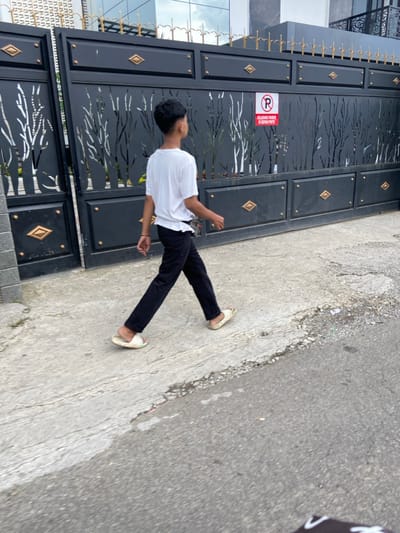 The image shows a young man walking on a paved road in front of a closed black gate with a "No Parking" sign. The gate is made of metal and has a decorative design of trees cut out in the middle. There are gold diamond-shaped accents on the gate. The man is wearing a white t-shirt, black pants, and white sandals. He is walking away from the camera and towards the right side of the image. The scene is located in Lembang, Indonesia, at coordinates -6.831 latitude and 107.607 longitude.