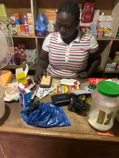 An overhead shot depicts a woman in Gbeleko, Nigeria, working at a counter in what appears to be a small pharmacy or dispensary. Shelves filled with various medications and health products are visible behind her. The counter is cluttered with items including containers of medications, boxes of drugs (TETRAKRIS, POLETRIM), bags, scissors, and miscellaneous papers. The woman, wearing a striped Fila shirt, is handling a sponge-like object. A partially visible electric fan sits in the lower left corner.